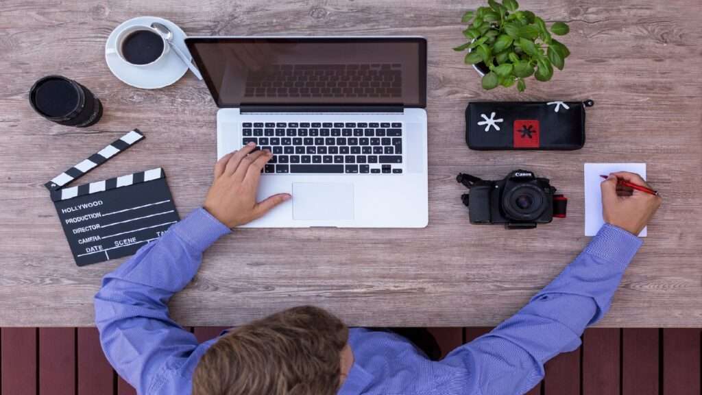 Mann sitzt an seinem Laptop und neben ihm befinden sich eine Filmklappe, ein Objektiv, eine Kamera und ein Kaffee.