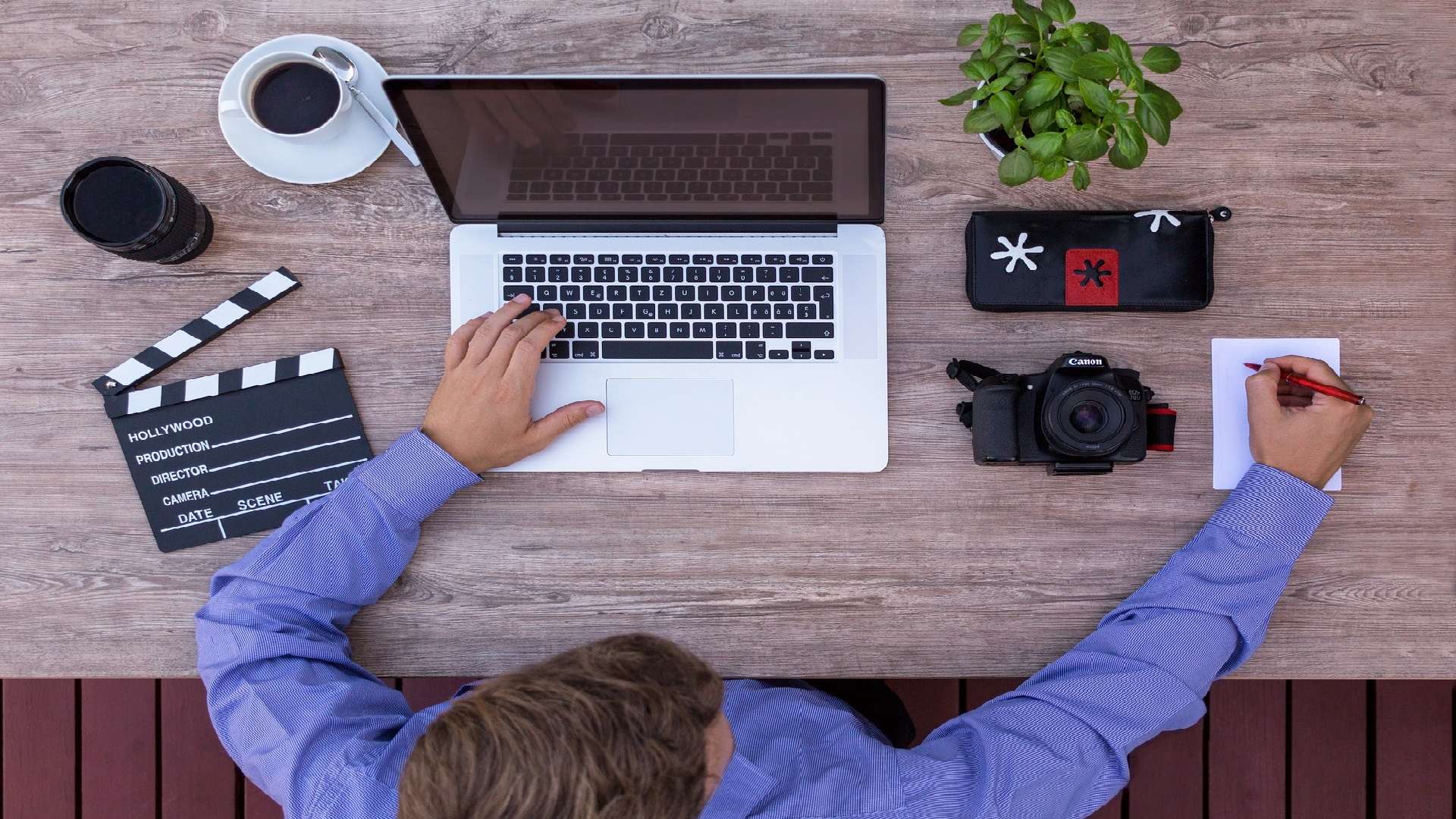 Mann sitzt an seinem Laptop und neben ihm befinden sich eine Filmklappe, ein Objektiv, eine Kamera und ein Kaffee.
