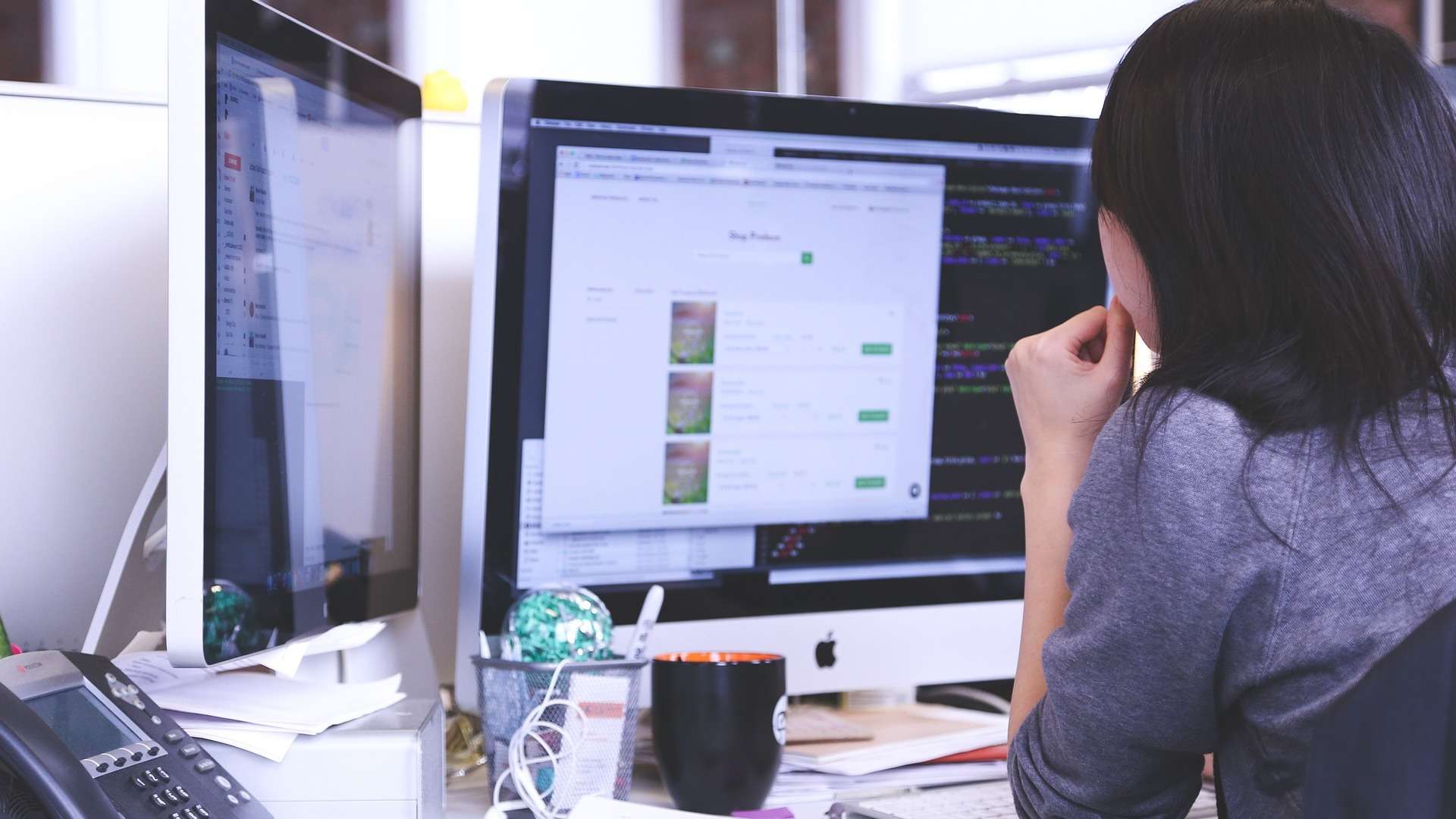 Ein Frau sitzt an einem Schreibtisch vor zwei iMacs und arbeitet