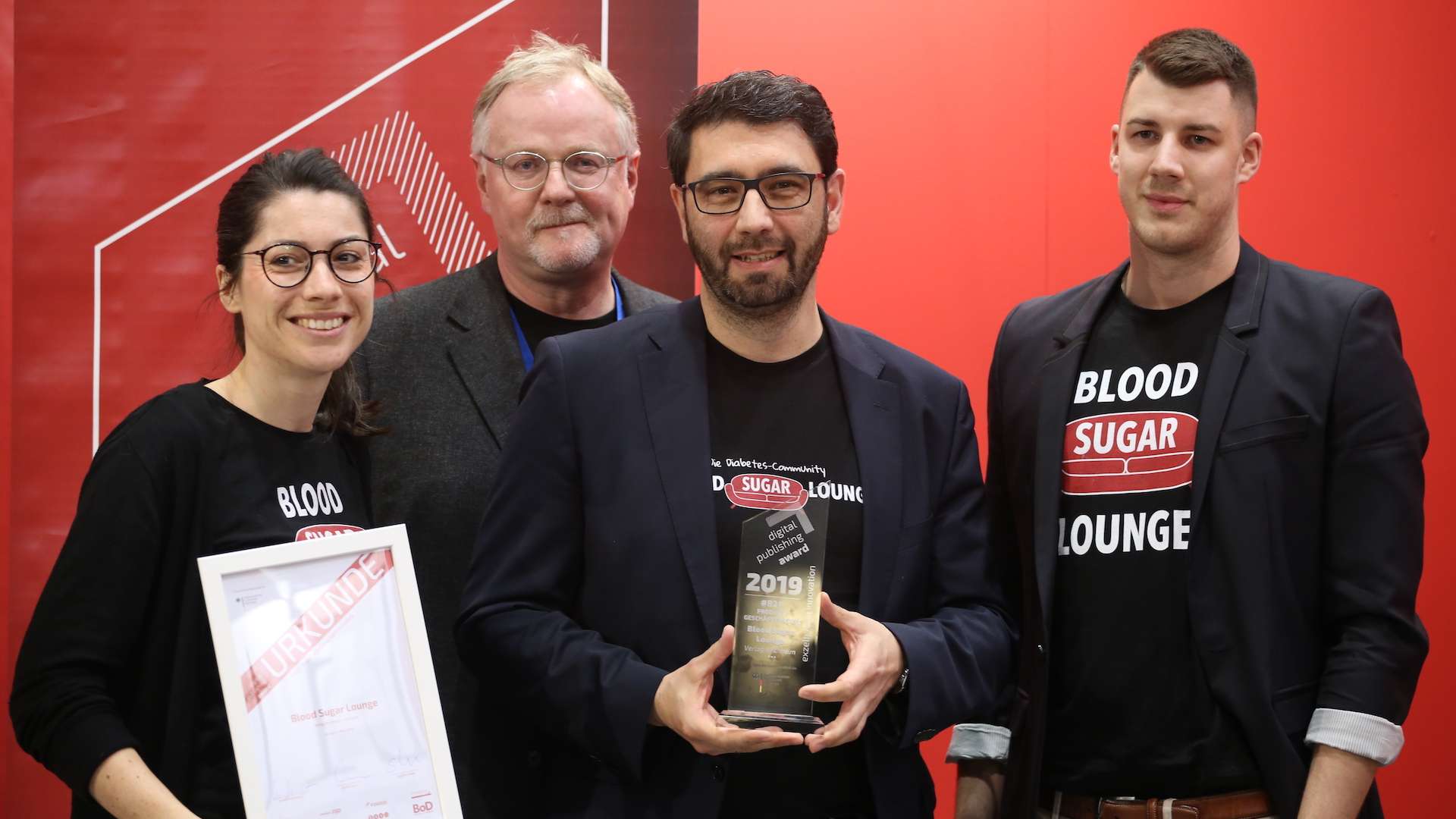 Vier Mitarbeiter der Blood Sugar Lounge stehen vor einem roten Hintergrund und präsentieren den gewonnen digital publishing award