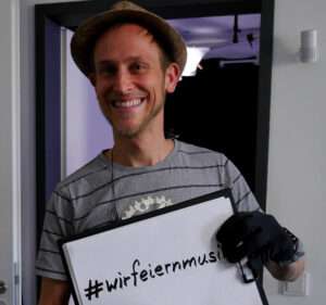 Bandmitglied beim Livestream vom Schlossgrabenfest@Home mit Schild "#wirfeiernmusik"