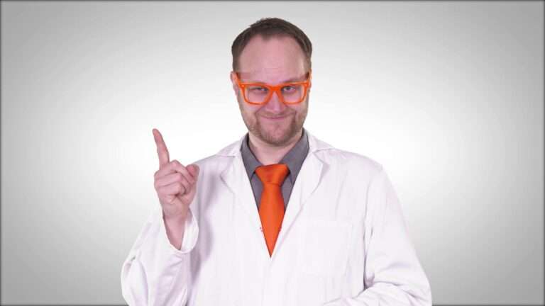 Ein Mann mit orangefarbener Brille und Krawatte in einem Laborkittel hebt den rechten Zeigefinger